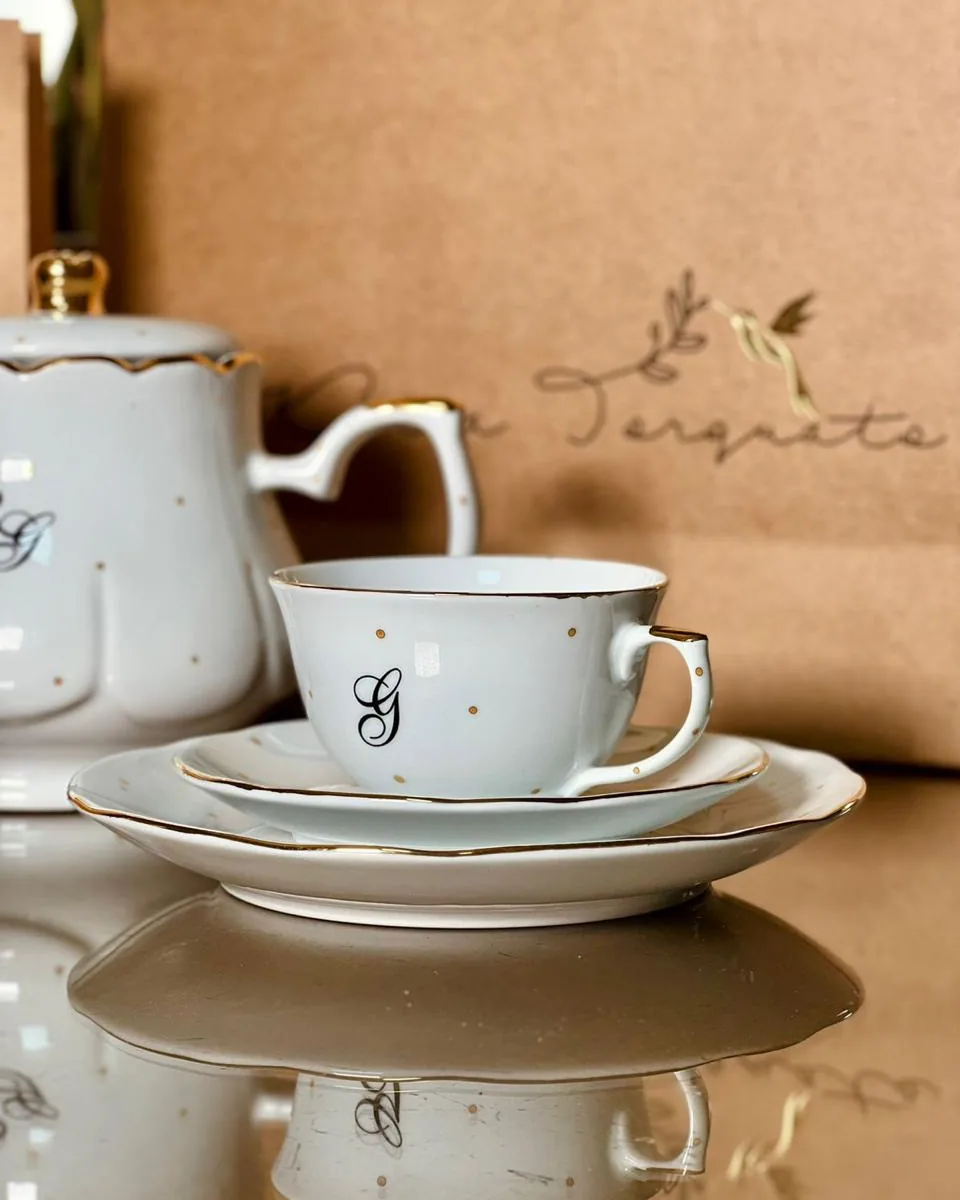 Xícara e pires de porcelana com monograma e friso de ouro, bule ao fundo, sacola kraft